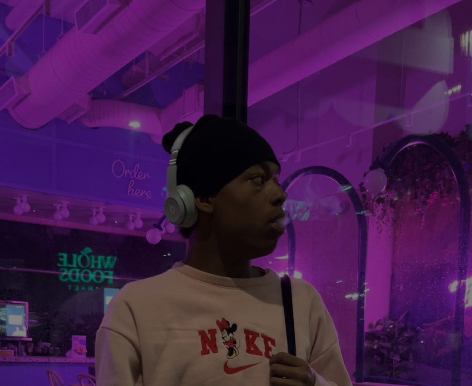 Close crop of the artist in headphones and a beanie against a purple-lit storefront window, mid-exhale.